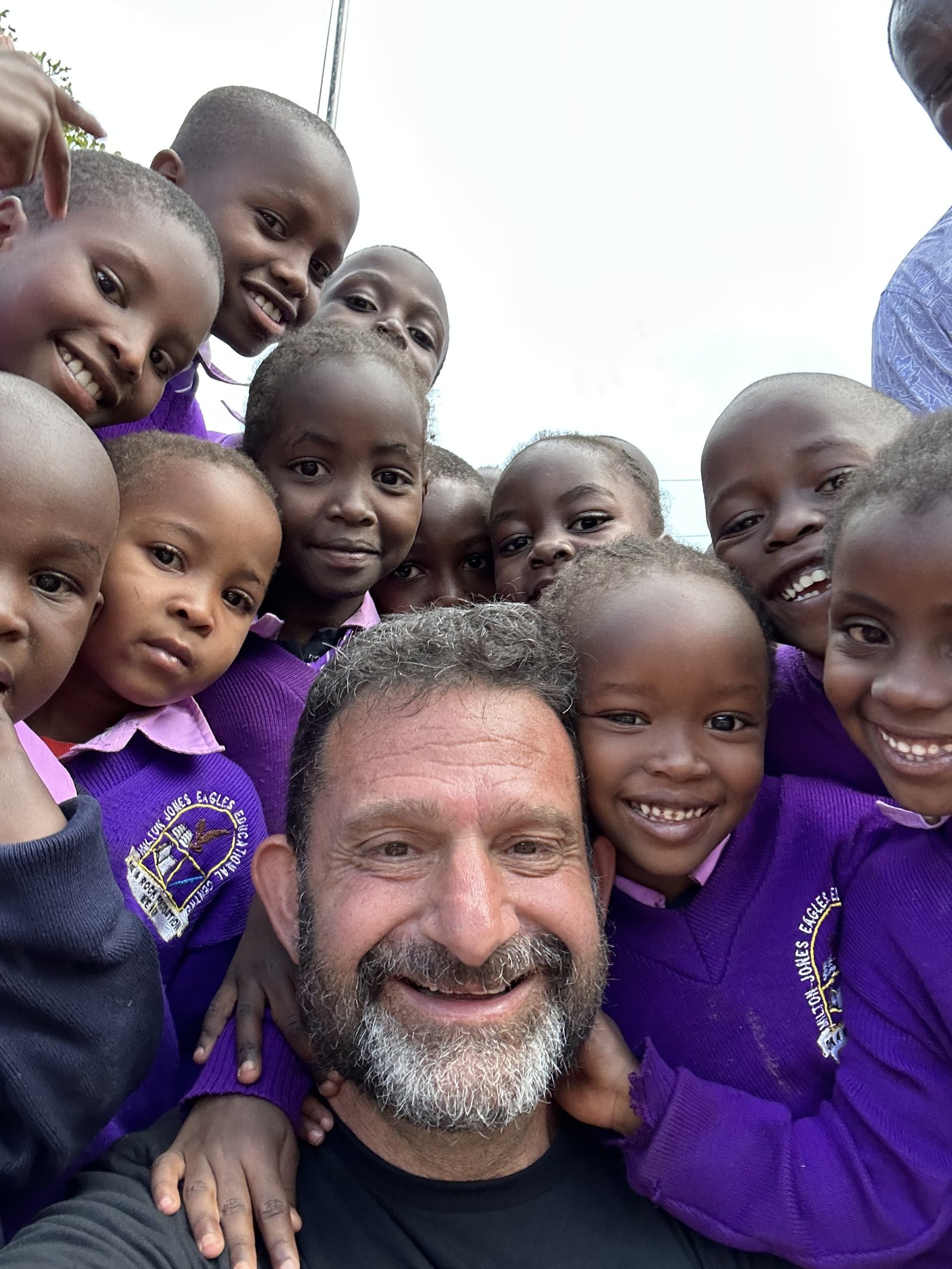 Darren Margolias taking a selfie surrounded by smiling children in Kenya in purple school uniforms