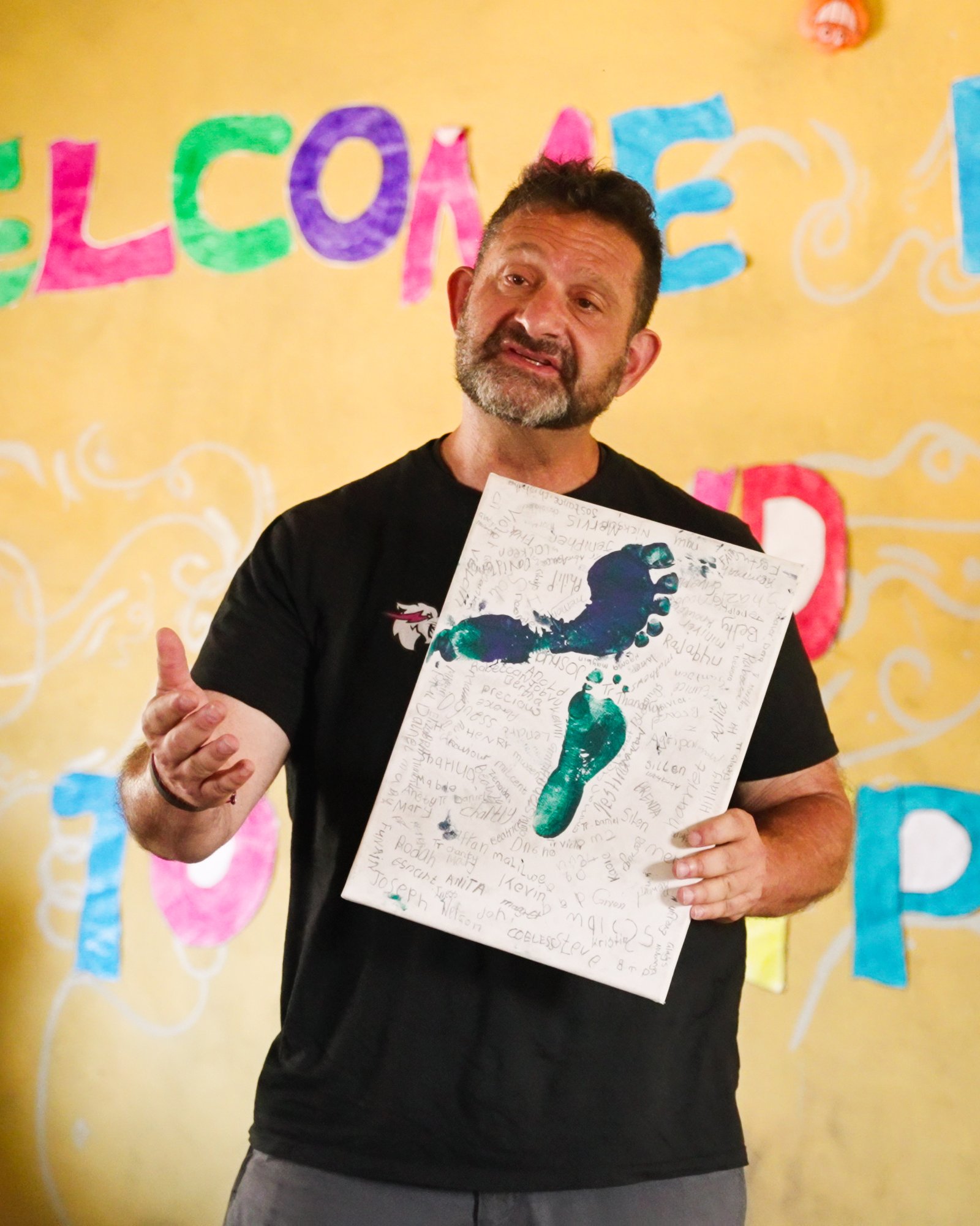 Darren Margolias holding a signed footprint poster in front of a colorful WELCOME mural in Zambia